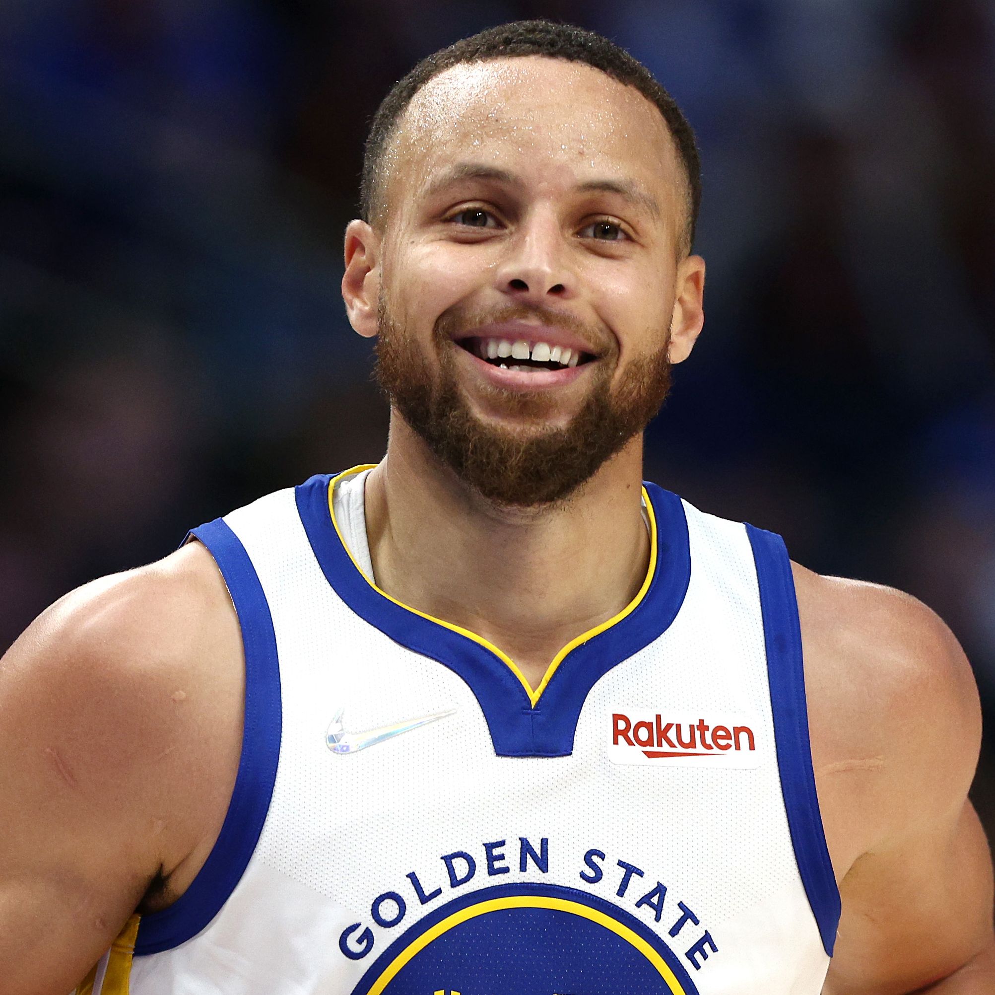 stephen curry smiles and looks past the camera, he wears a white golden state warriors jersey with blue and yellow accents